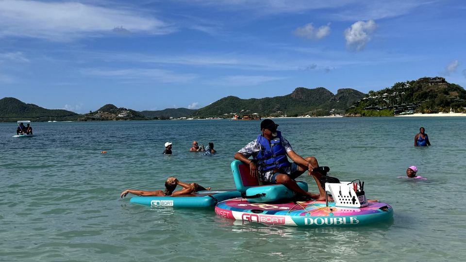 A guest drives a pink and turquoise GoBoat on calm turquoise water off Antigua's west coast, with a second guest relaxing on the front float and green hills behind.