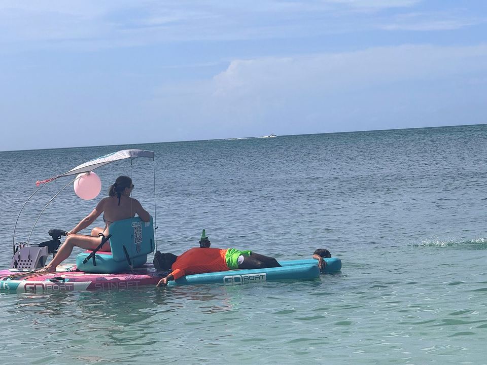 A woman steers a GoBoat under its sun shade while her partner stretches out on the front float on a calm sea.