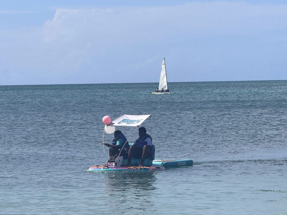 Two friends cruise side by side on a GoBoat with a sun shade and pink balloon, with a white sailboat in the distance.