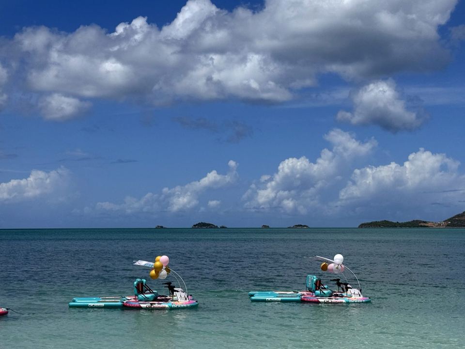 Two GoBoats with balloons glide across clear water under a bright blue sky with big white clouds.