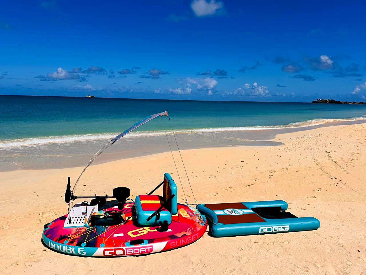 An empty pink and turquoise GoBoat Double with its sun-shade pole and side float on white sand beside clear blue water.